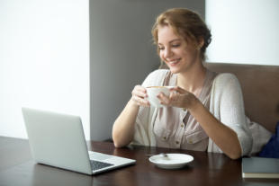 Half-length portrait of young beautiful female working on laptop in coffee shop. Woman sitting with cup of coffee and looking at laptop screen. Attractive model using computer in cafe