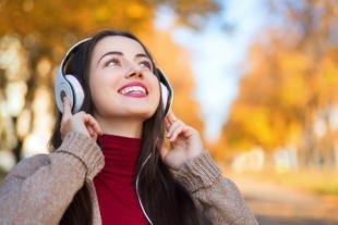 Young happy smiling brunette woman with headphones outdoors on autumn day. Girl listening music in headphones in autumn park. Portrait of woman at outdoor with headphones. Autumn portrait of happy girl. Copy space